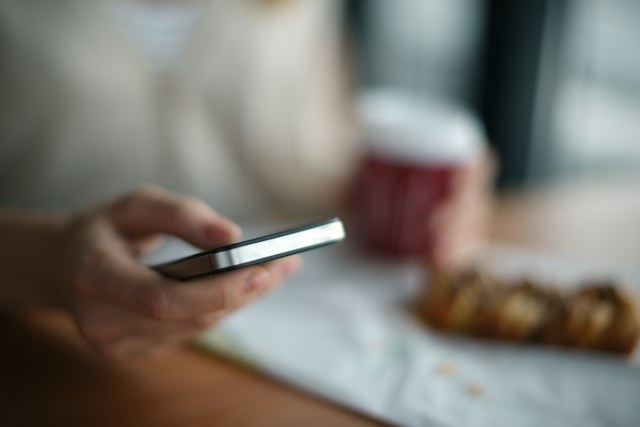 Ruke drže mobilni telefon dok prst dodiruje ekran pored šolje