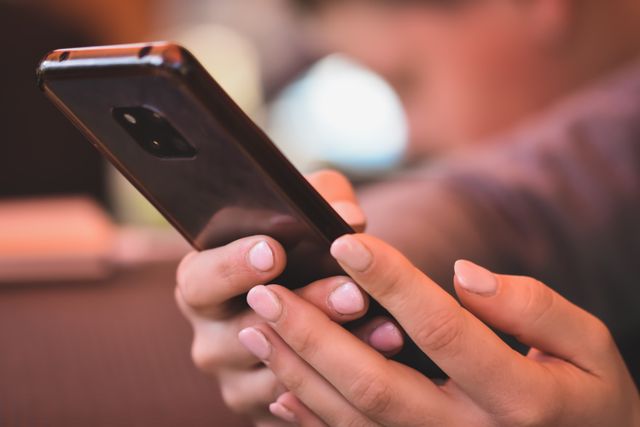 Ruke drže mobilni telefon sa tamnim kućištem i svetlo roze noktima